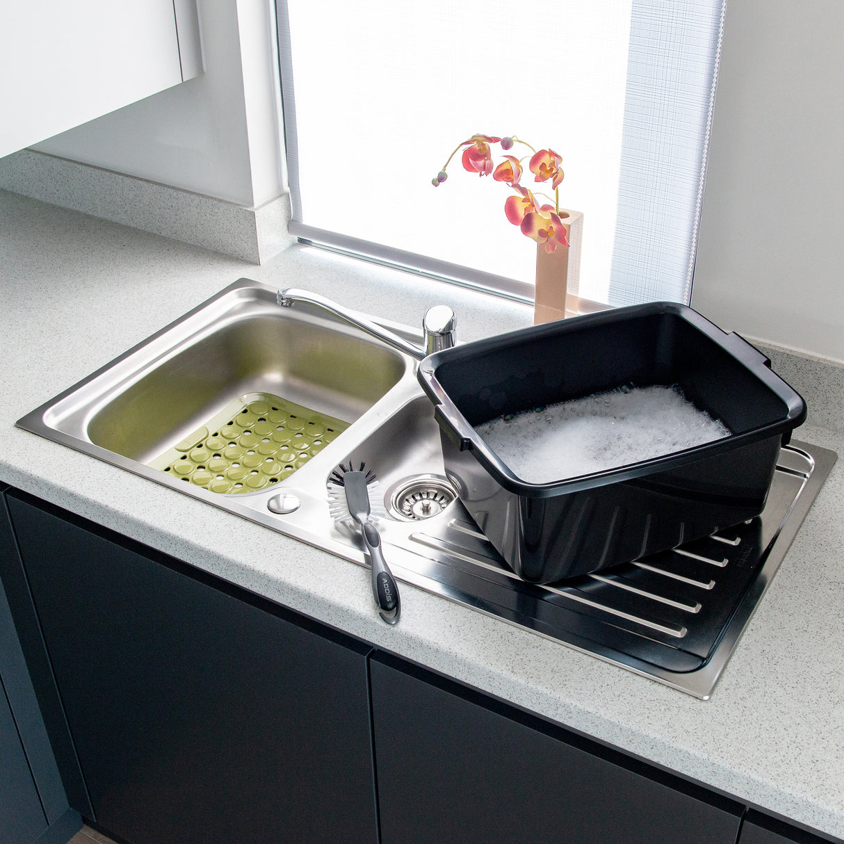 Kitchen sink with a large black washing up bowl filled with soapy water on a light grey countertop, alongside other sink accessories including a dish brush and sink liner.