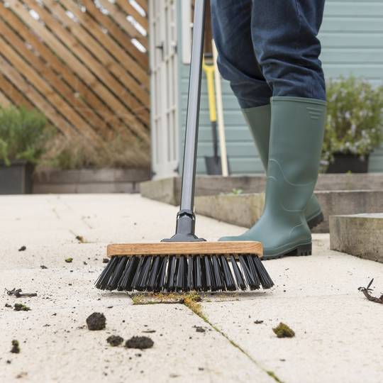 Person in green wellington boots using a stiff bristled broom to clean a patio surface.