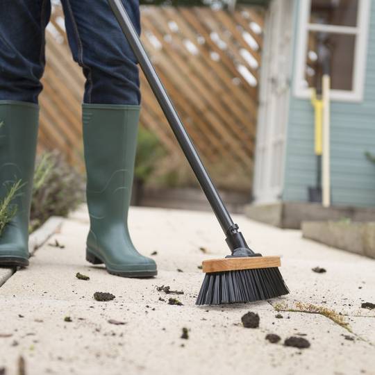 Person in green wellington boots using a stiff bristled broom on a concrete surface.