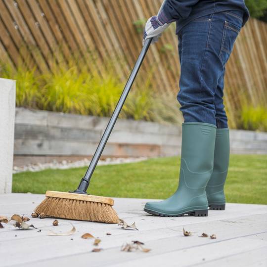 Person in green wellington boots using a broom in a garden setting to sweep leaves.