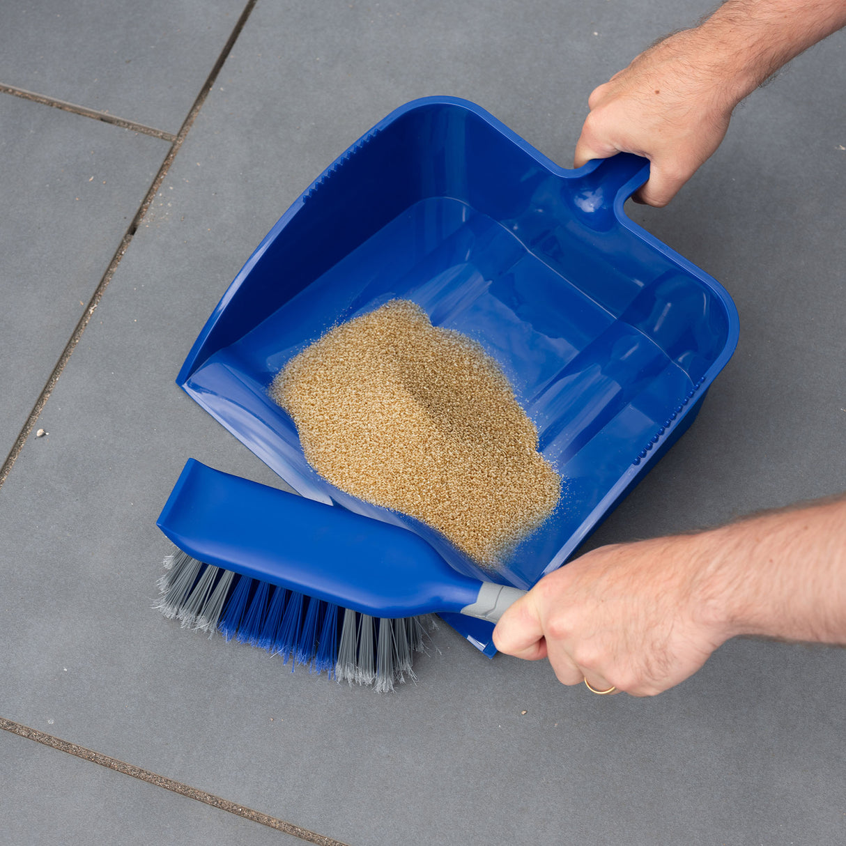 Blue dustpan and brush being used to sweep sand from a grey patio