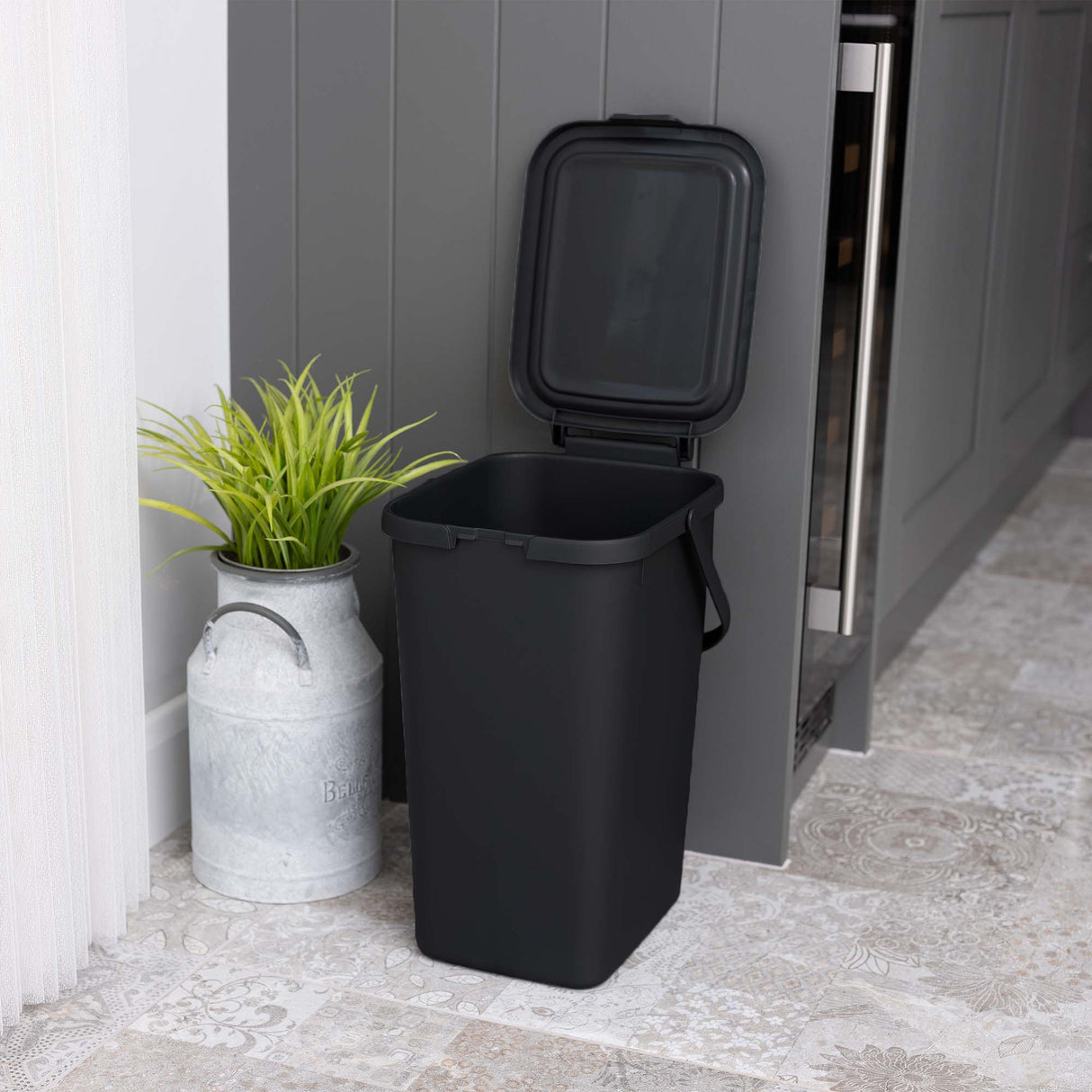 Small black utility caddy with lid open ready for recycling, next to a grey cupboard with a plant in a metal pot.