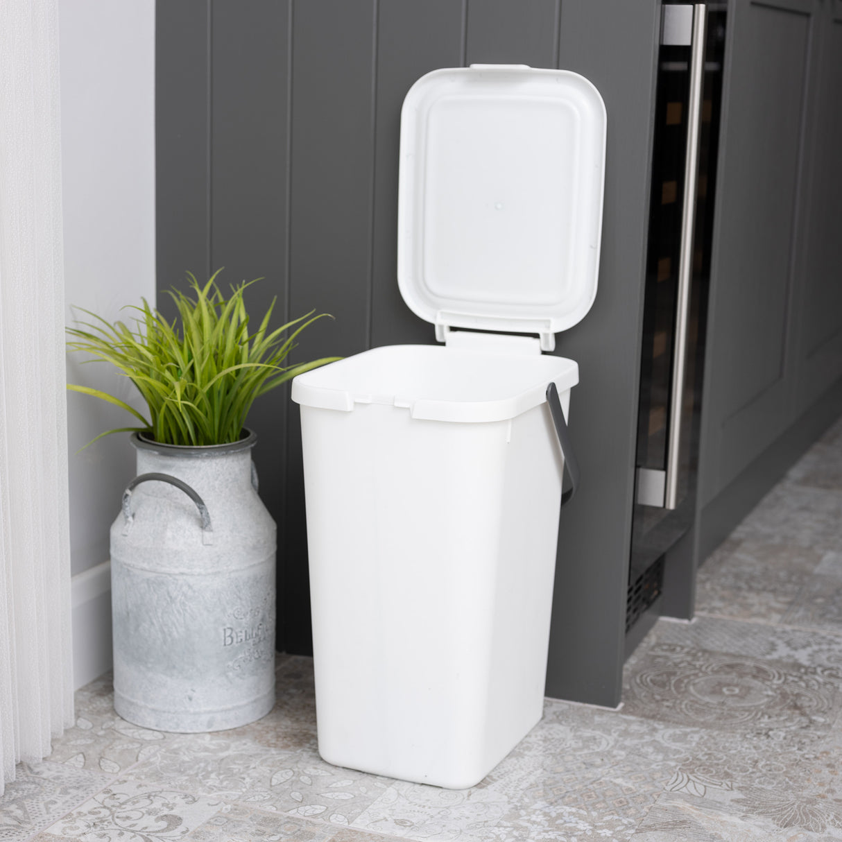 White utility caddy with lid open waiting to be used for recycling, next to a grey cupboard and plant in metal pot.