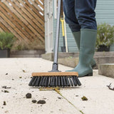 Person in green wellington boots using a stiff bristled broom to clean a patio surface.