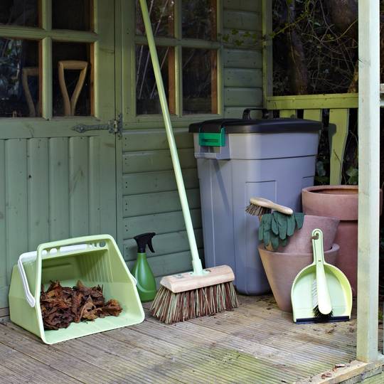 Gardening tools and accessories including a large wooden broom on a wooden deck next to a green shed.